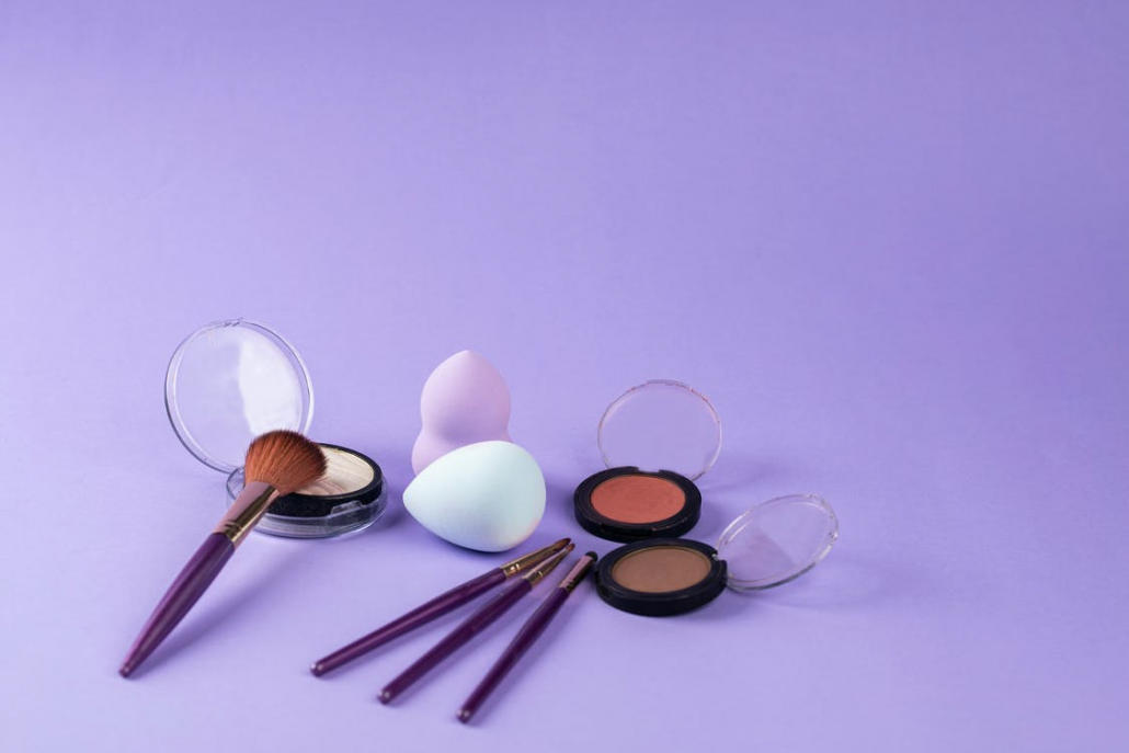 Some Makeup Brushes, Two Sponges, and Some Compact Makeup on a Purple Surface